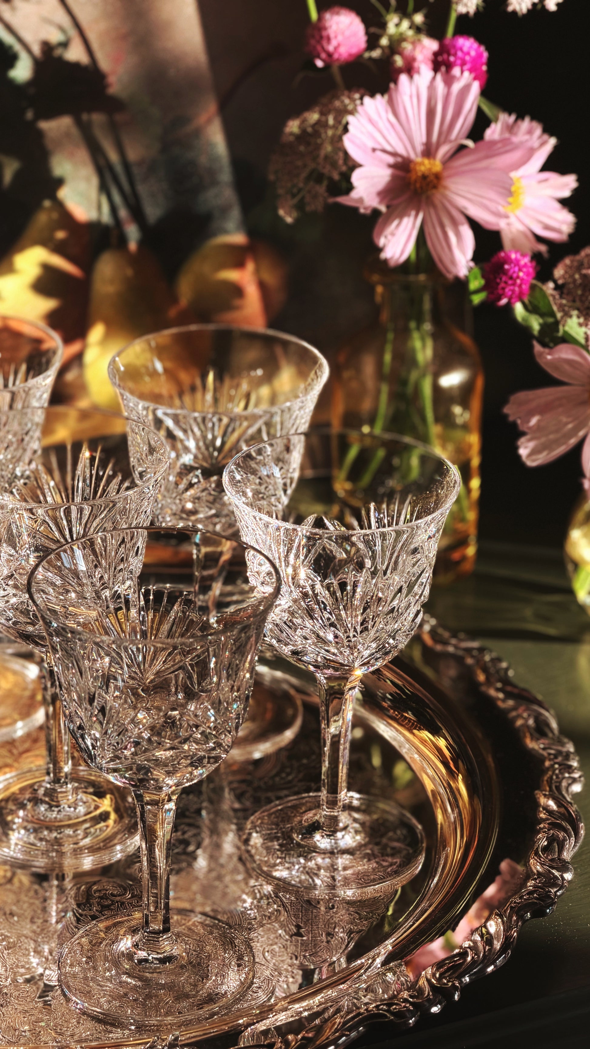 A set of clear cut-crystal wine glasses arranged on an ornate silver tray, with pink flowers and a warm-toned bottle in the softly blurred background.