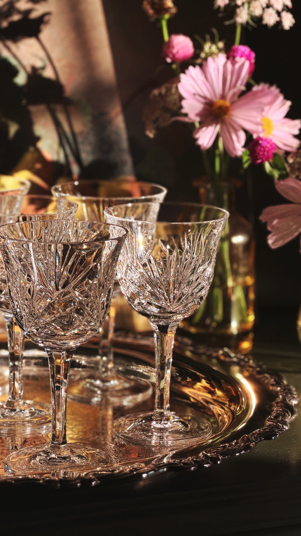 A group of clear cut-crystal wine glasses arranged on an ornate round silver tray, with a blurred background of pink flowers and warm ambient lighting.