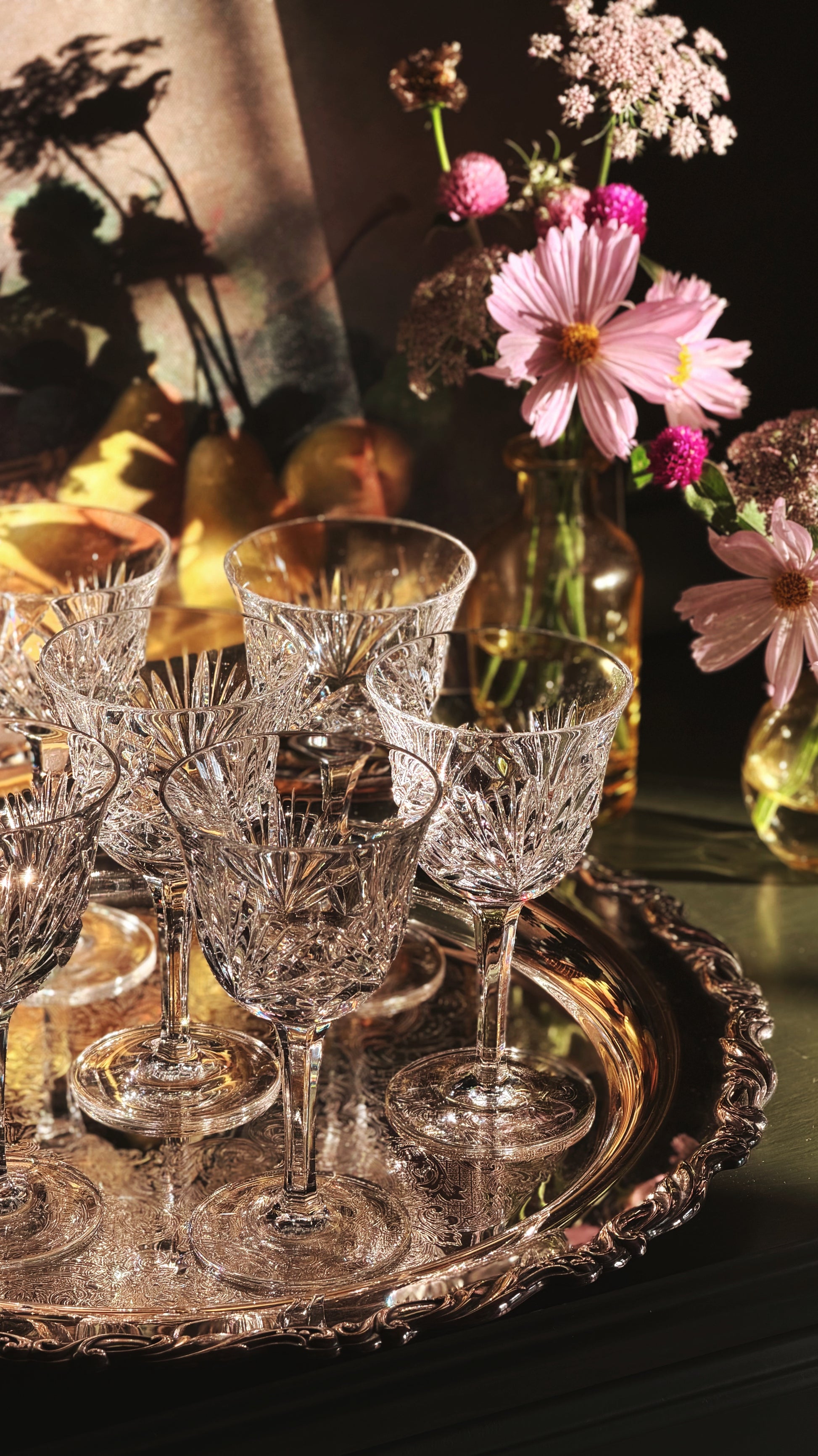 A set of clear cut-crystal wine glasses arranged on an ornate silver tray, styled with pink flowers in glass bottles against a dark, warm-toned background.
