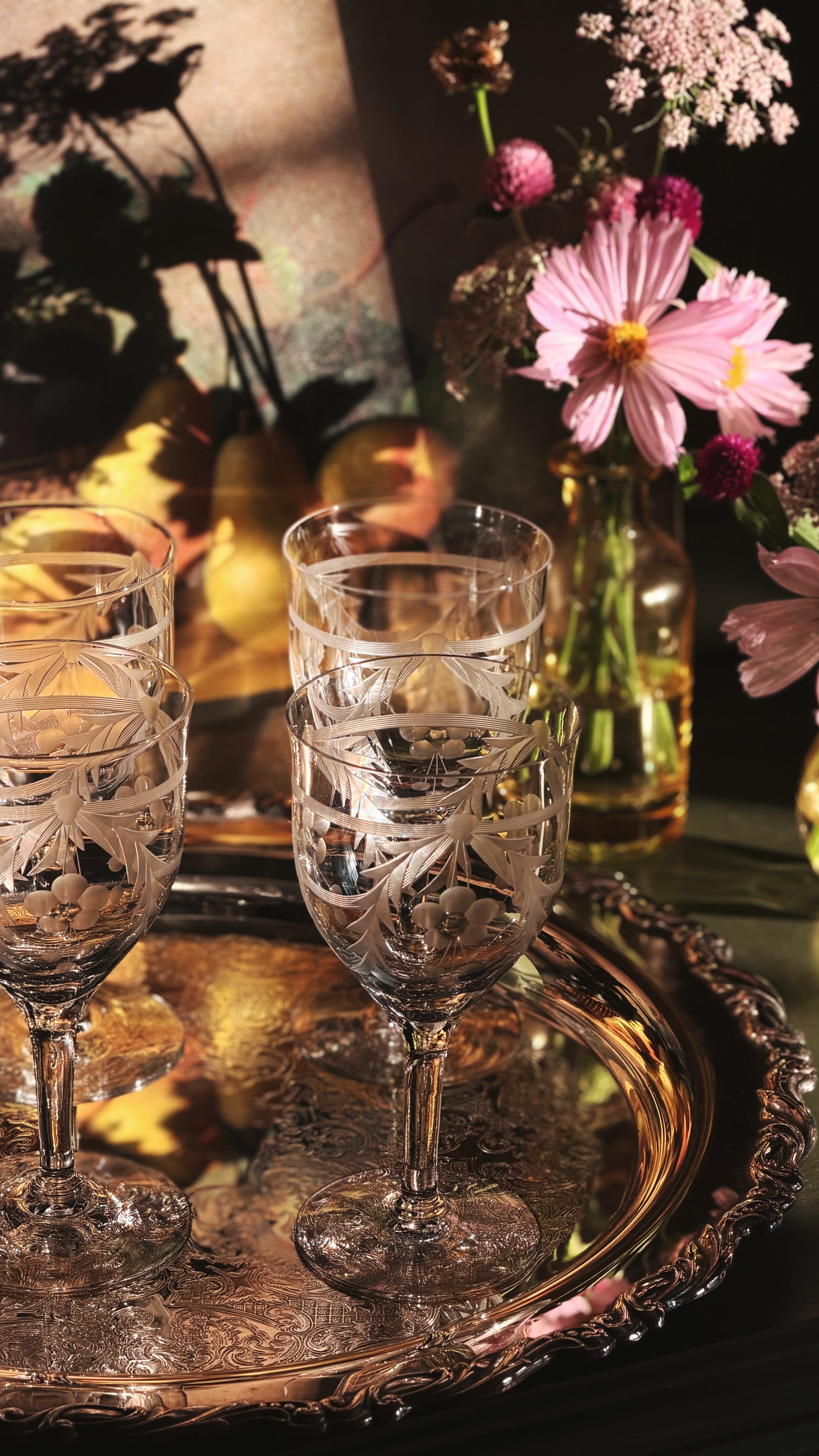 Two clear cut-crystal wine glasses stacked on an ornate round silver tray, with a blurred floral arrangement in the background under warm sunlight.