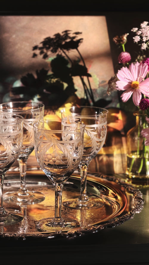 A set of clear cut-crystal wine glasses arranged on a round silver tray, with warm candlelight and a floral arrangement in the background.