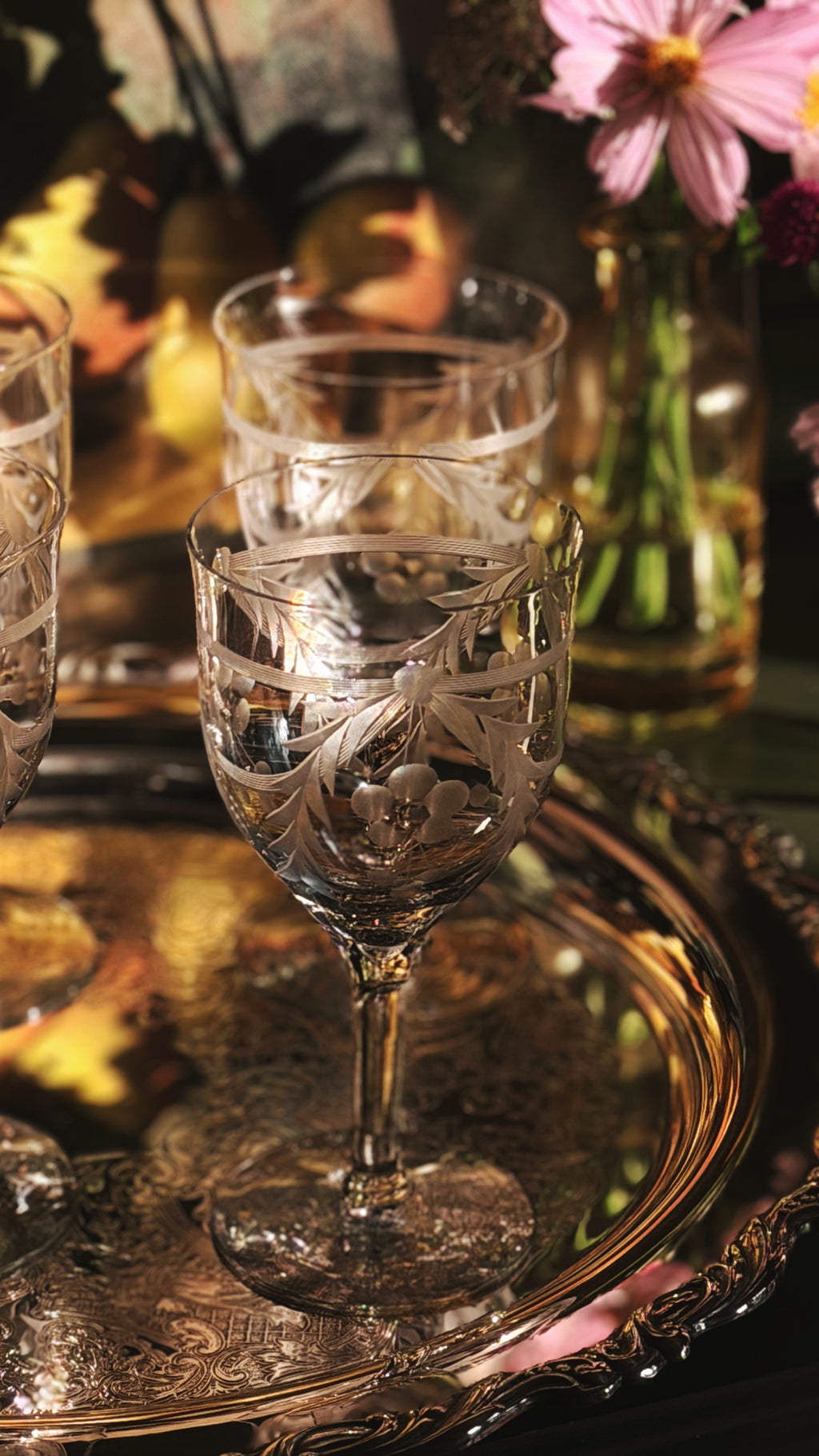 A close-up of a clear etched glass goblet with a stem, placed on an ornate gold tray, with other matching glasses and pink flowers in the background.