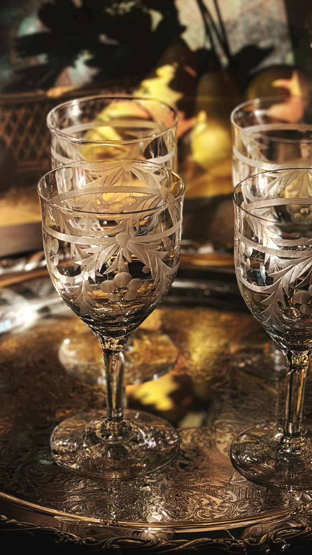 Three clear etched crystal wine glasses with stems, arranged on an ornate reflective silver tray with warm golden background lighting.