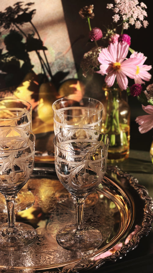 Three clear etched crystal wine glasses arranged on an ornate silver tray, with a warm floral and golden background in a moody styled setting.