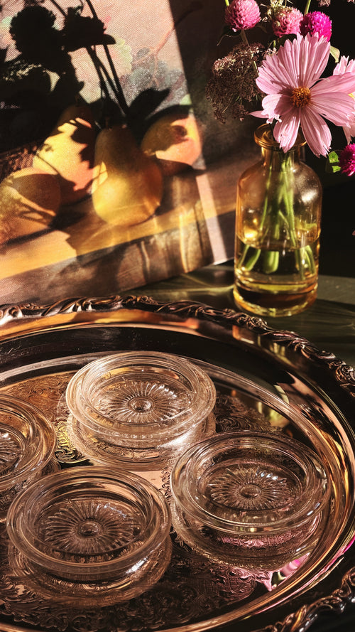 Four clear glass coaster with embossed floral patterns arranged on an ornate silver tray, with a glass vase of pink flowers in the background under warm directional light.