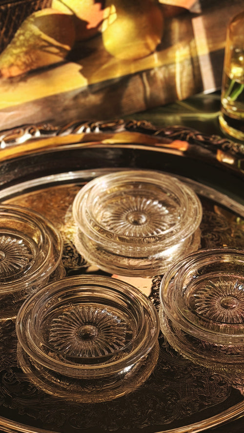 Four clear glass coasters with a sunburst textured pattern, arranged on a decorative silver-toned metal tray with gold trim.