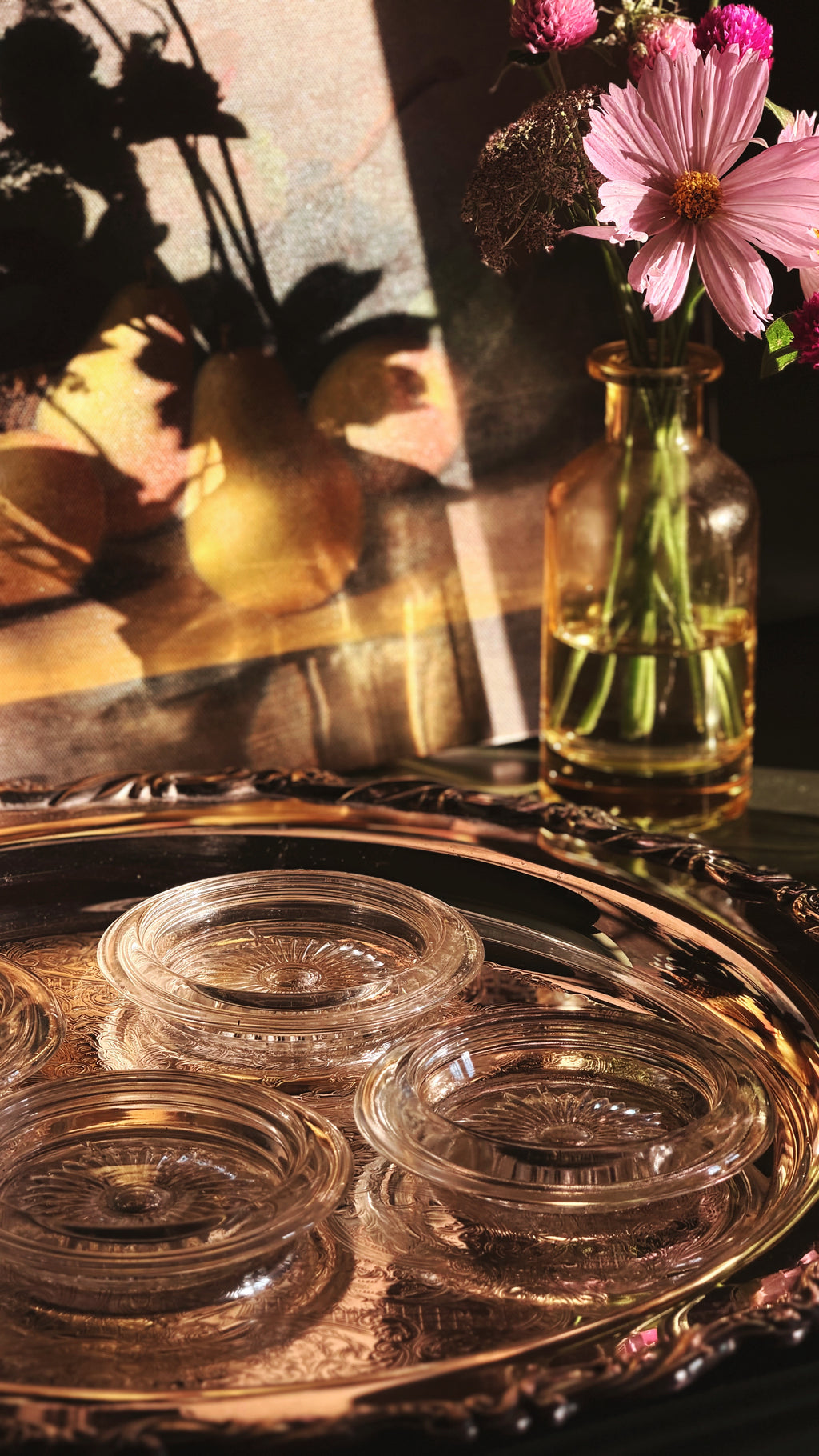 Four clear glass coasters arranged on an ornate round silver tray, with a small glass vase of pink flowers in the background under dramatic warm lighting.