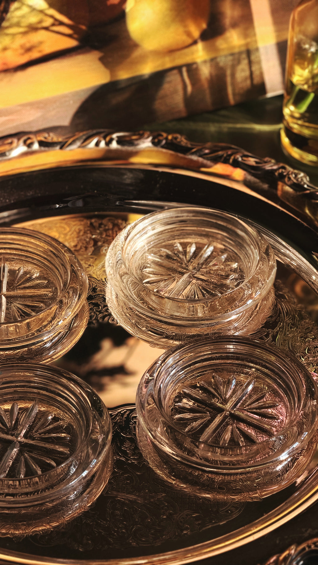 Four clear glass coasters with starburst patterns, arranged on a dark decorative tray with warm golden lighting.