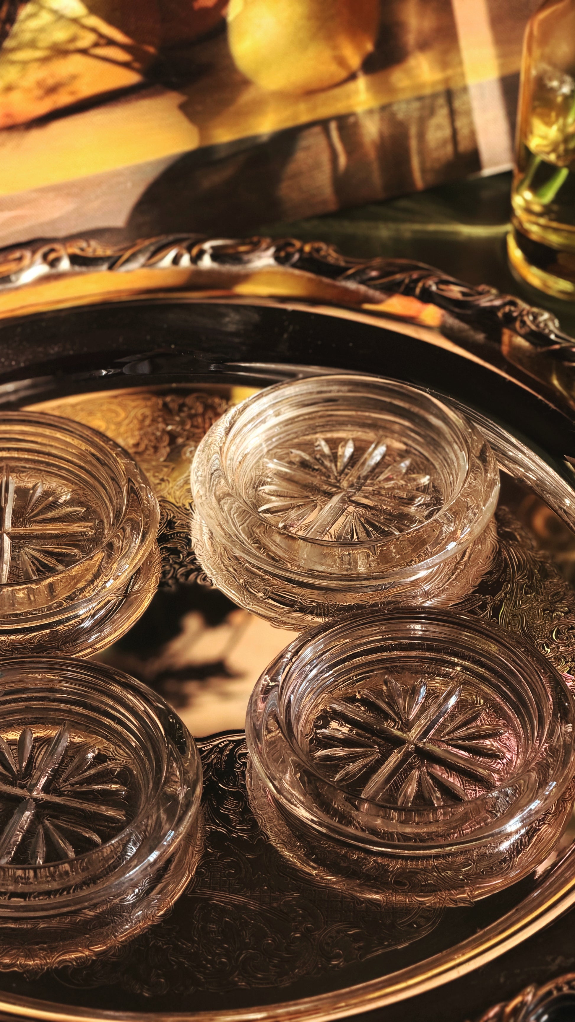 Four clear glass coasters with starburst patterns, arranged on a dark decorative tray with warm golden lighting.
