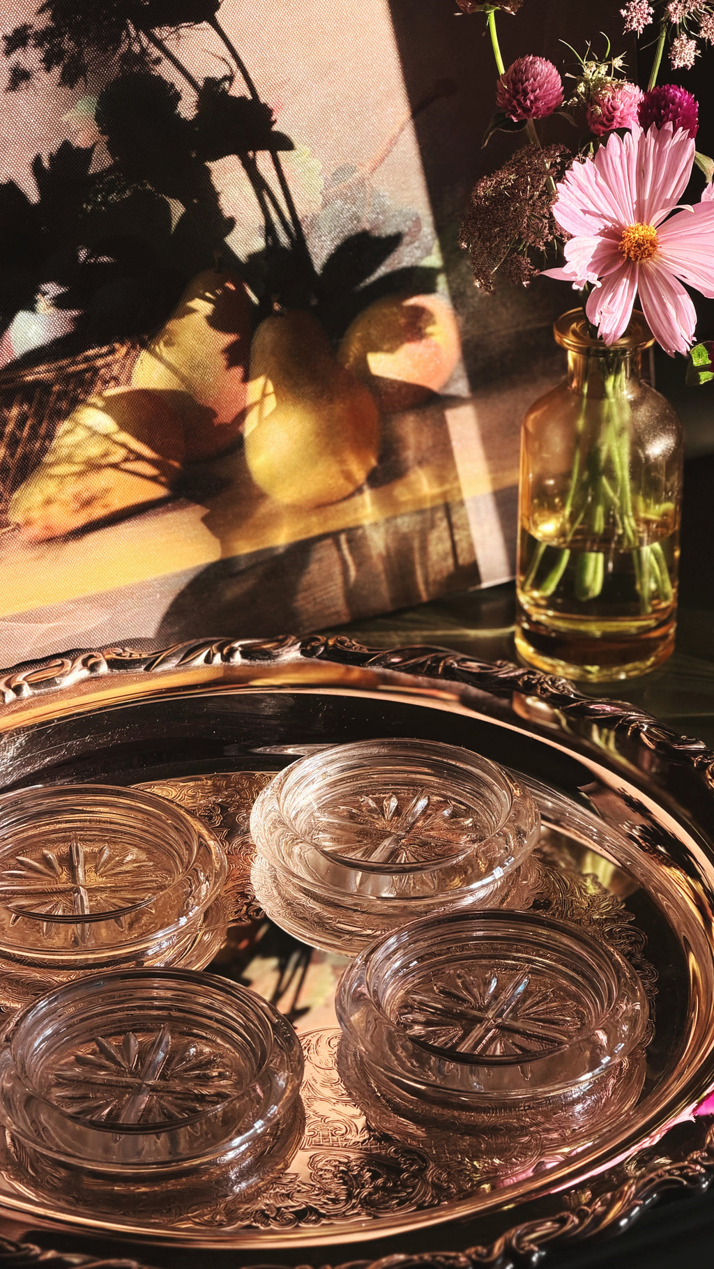 Four clear glass coasters with embossed starburst patterns arranged on an ornate silver tray, with a glass vase of pink flowers and a warm-toned still life painting in the background.