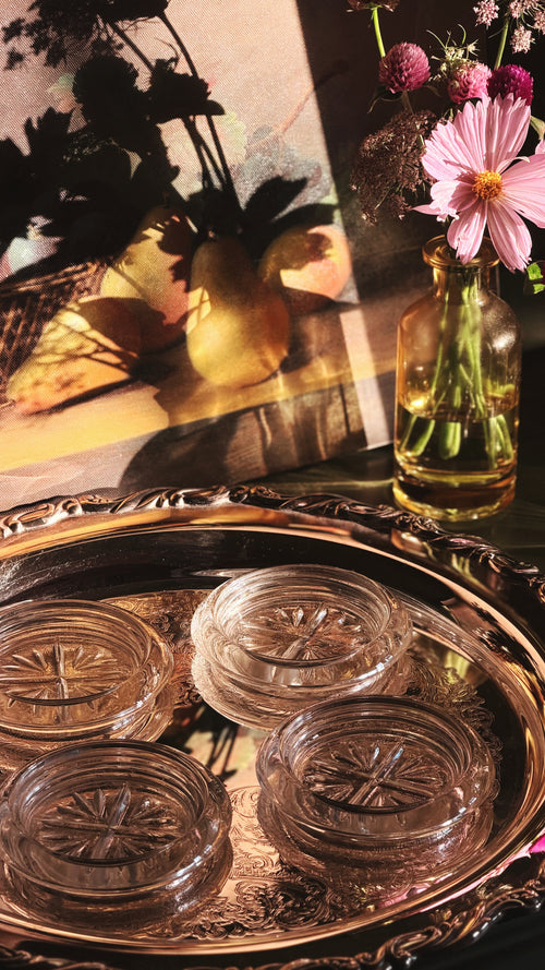 Four clear glass coasters with embossed starburst patterns arranged on an ornate silver tray, with a glass vase of pink flowers and a warm-toned still life painting in the background.
