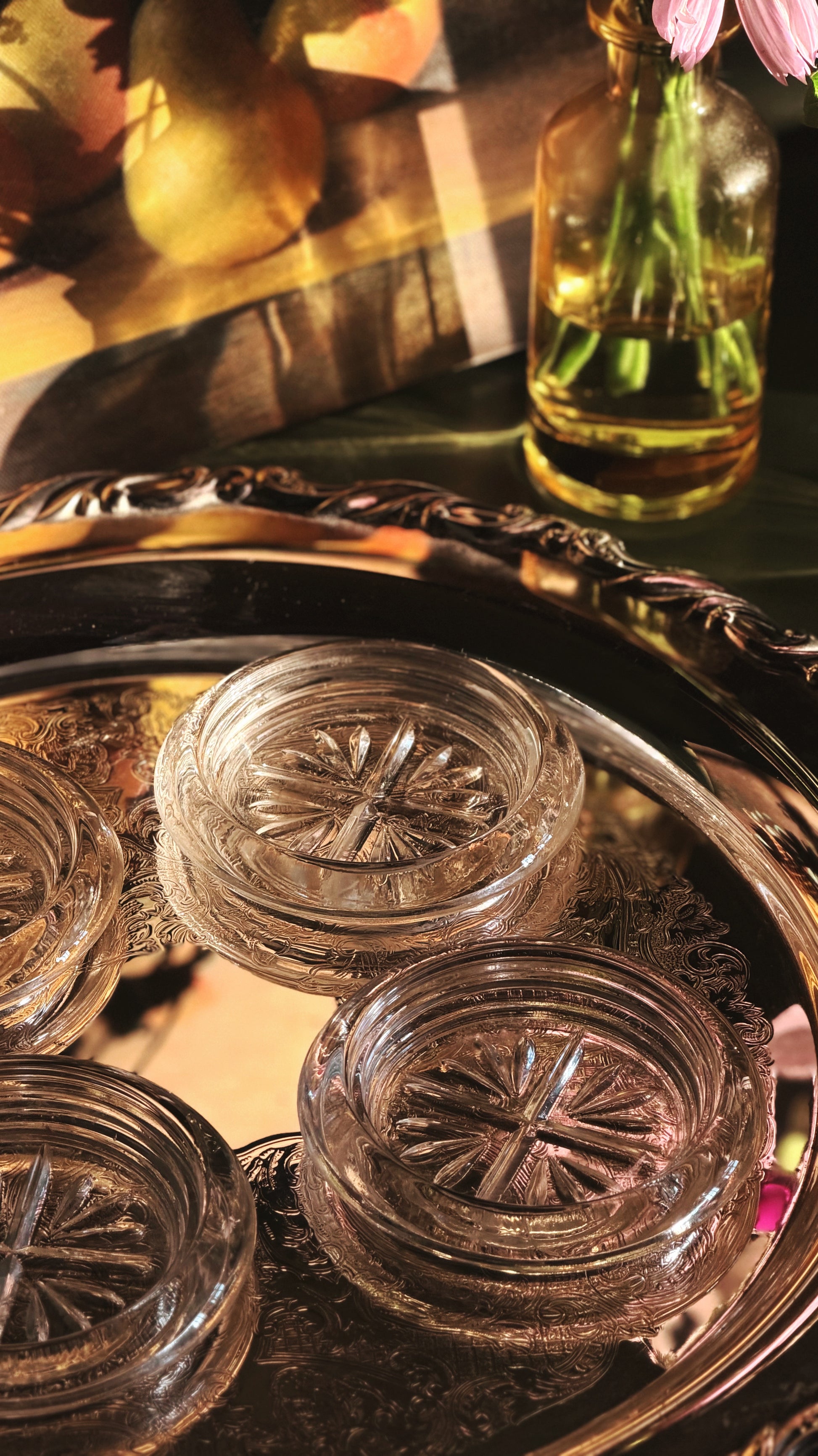 Clear glass coasters with embossed starburst patterns arranged on an ornate silver tray, with a warm-toned decorative background and a small glass bottle of flowers in the upper right.