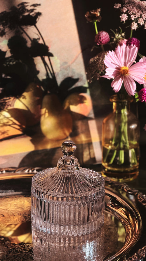 A clear ribbed glass jar with a matching lid and finial, styled on an ornate gold tray with pink flowers and a warm-toned background.
