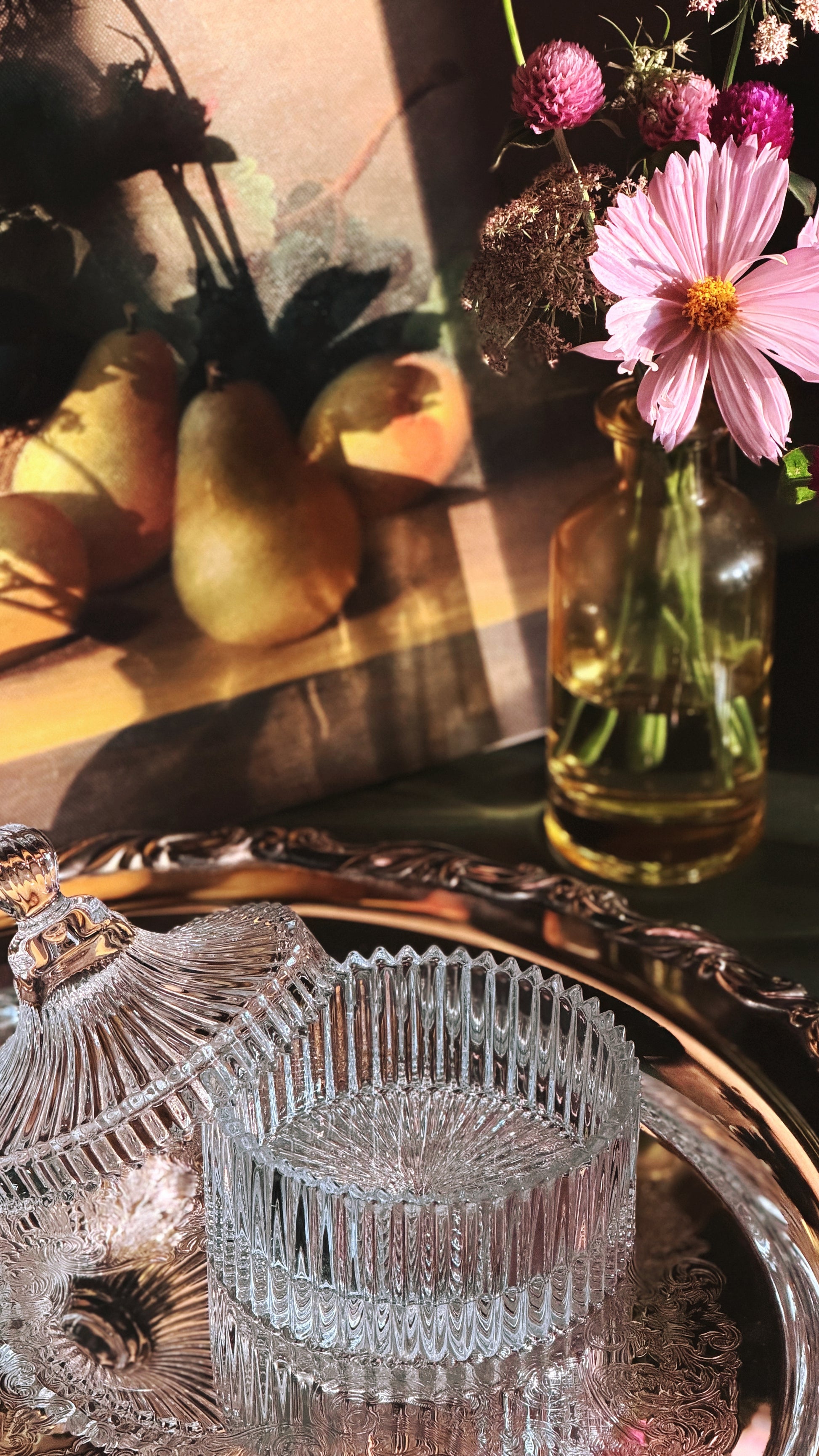 A styled still life scene with a clear ribbed glass jar and matching glass lid on an ornate silver tray, alongside a pink daisy in a glass bottle and pears in warm sunlight.
