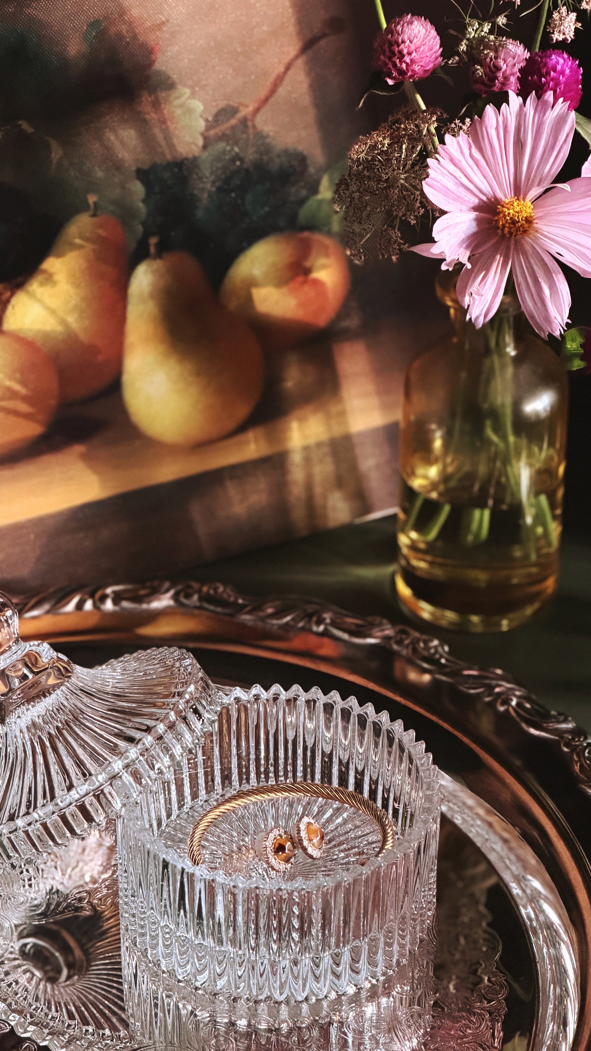 A close-up still life showing a faceted clear glass bowl with a gold-toned rim on an ornate silver tray, alongside a glass vase with pink flowers. In the softly blurred background, pears and a painting are visible.