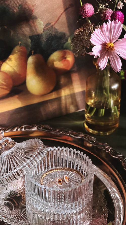 A close-up still life showing a faceted clear glass bowl with a gold-toned rim on an ornate silver tray, alongside a glass vase with pink flowers. In the softly blurred background, pears and a painting are visible.