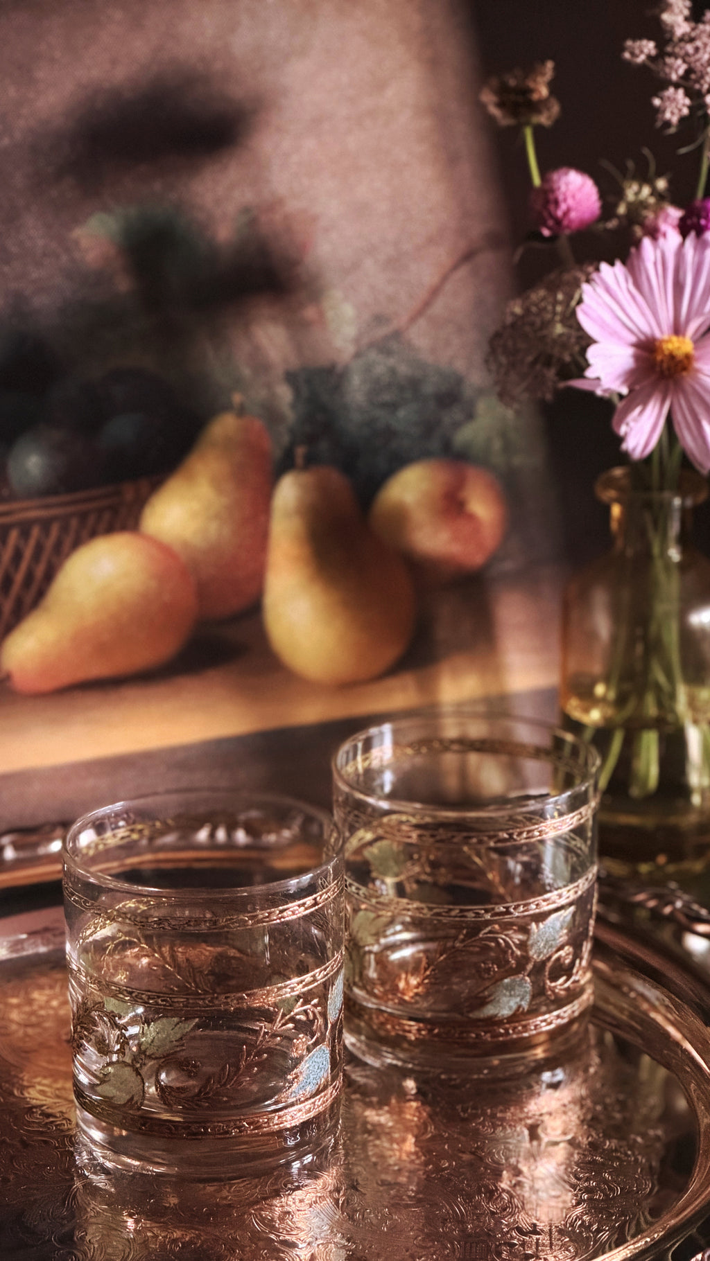 Two ornate clear glass tumblers with etched floral patterns and gold-toned bands, set on a reflective copper tray. The background is a warm, softly blurred still life with pears and a vase of pink flowers.