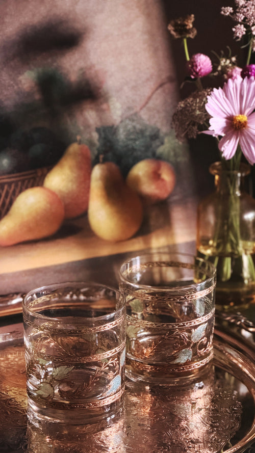 Two clear glass tumblers with embossed floral patterns, resting on a reflective metallic tray. The background is a softly blurred still life featuring pears, a pink flower in a glass bottle, and warm ambient lighting.