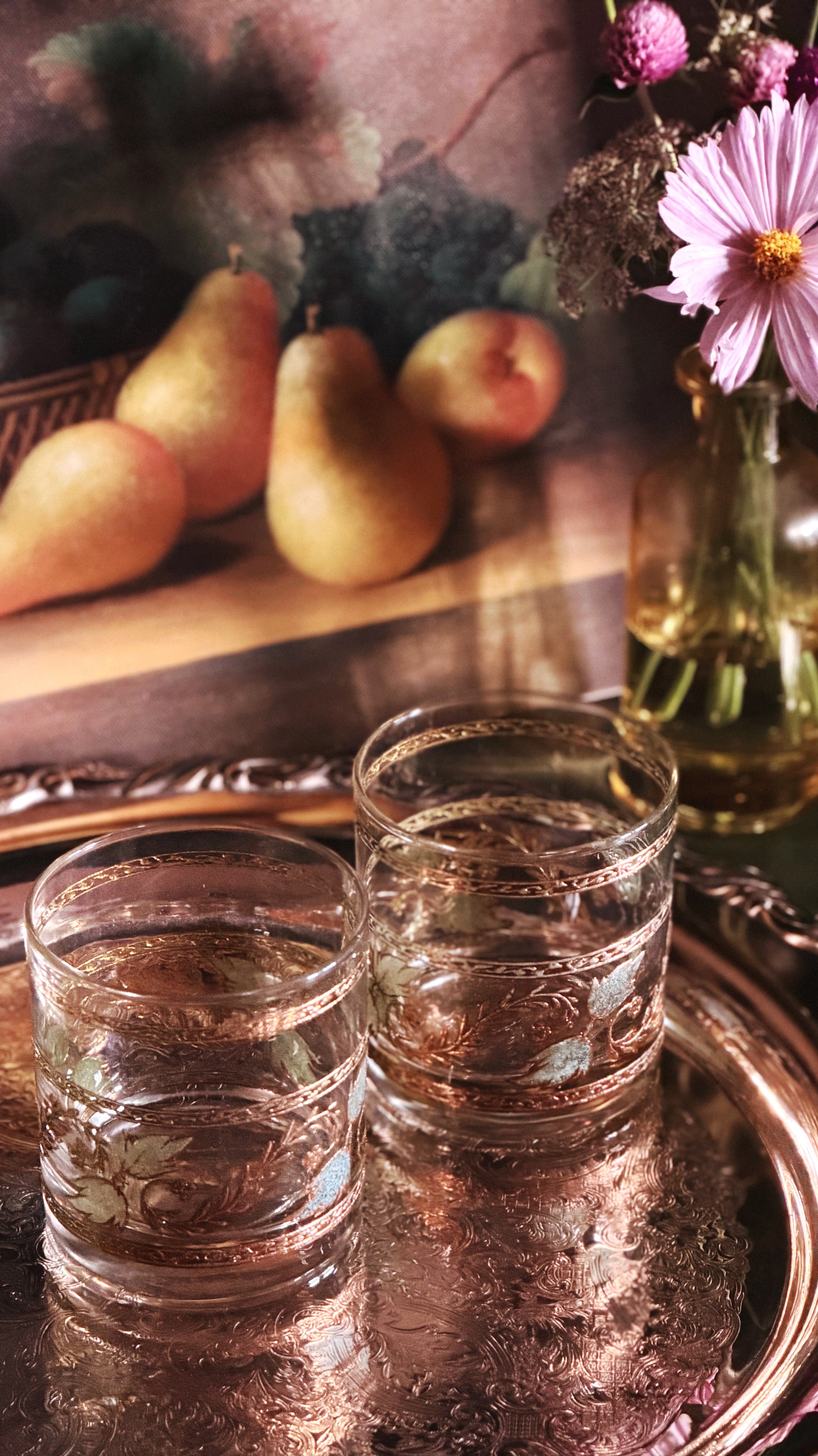 Two clear glass tumblers with etched floral patterns sit on a reflective copper tray, with a blurred still life background showing pears, a pink flower, and a glass vase.