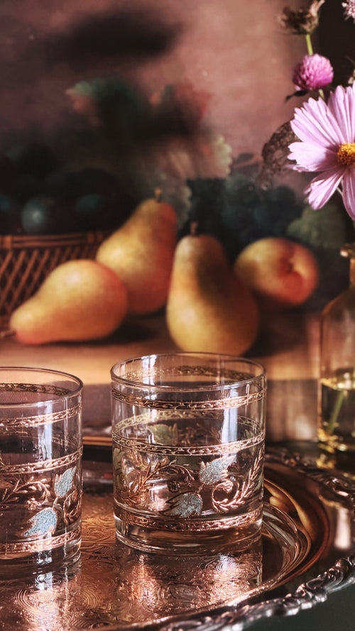 Two ornate clear glass rocks with etched floral patterns sit on a reflective copper tray, set against a warm, rustic still life background with pears, flowers, and soft shadows.