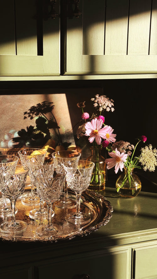 A styled interior scene with a silver tray holding several clear cut-crystal wine glasses on a green cabinet, alongside pink and white flowers in glass vases under dramatic warm sunlight and shadows.