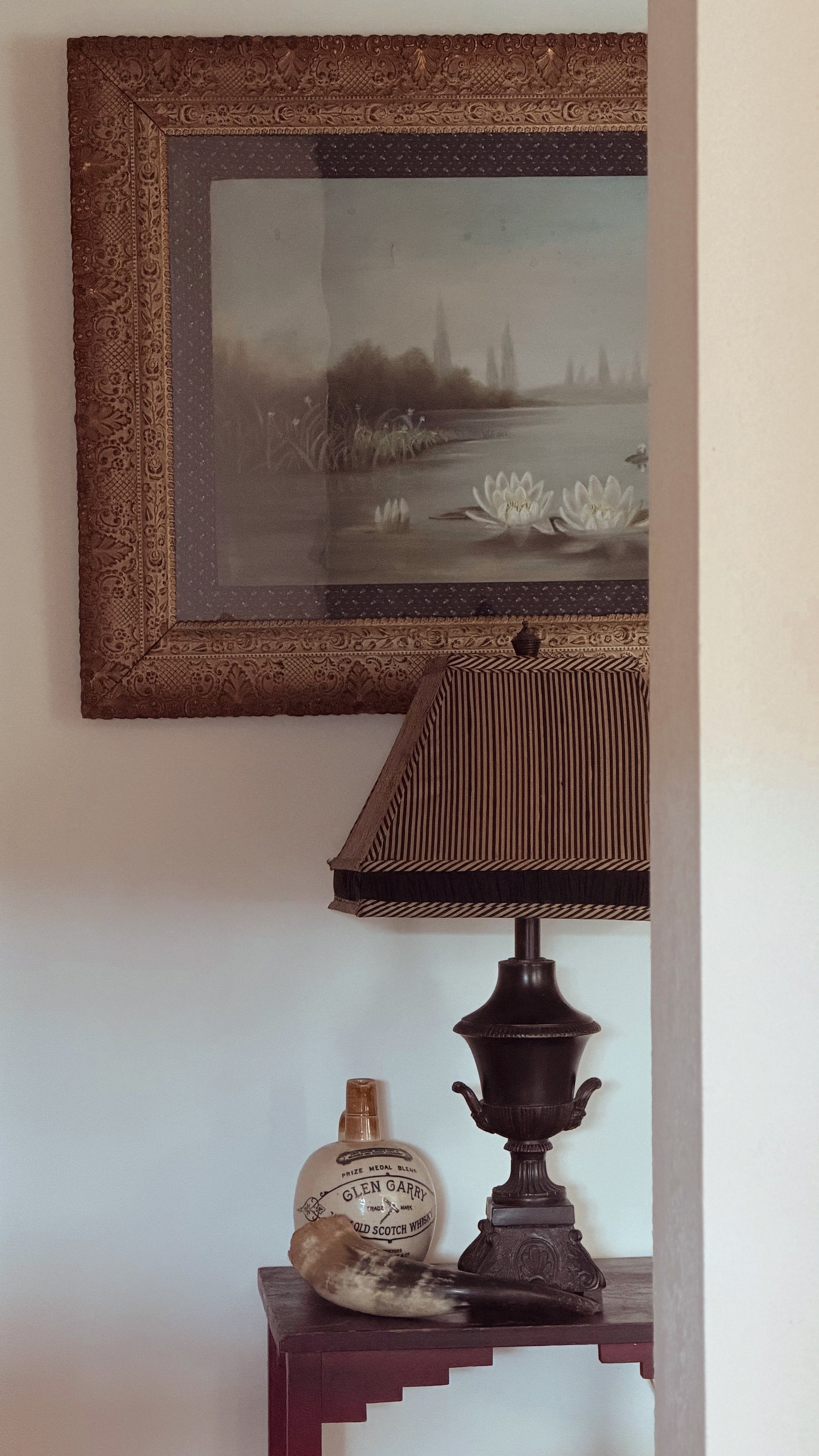 A framed landscape painting with a detailed brown ornate frame hangs on a light beige wall above a dark wood side table. The table holds a dark brown table lamp with a textured shade, a small decorative ceramic bottle, and a fur trim piece.