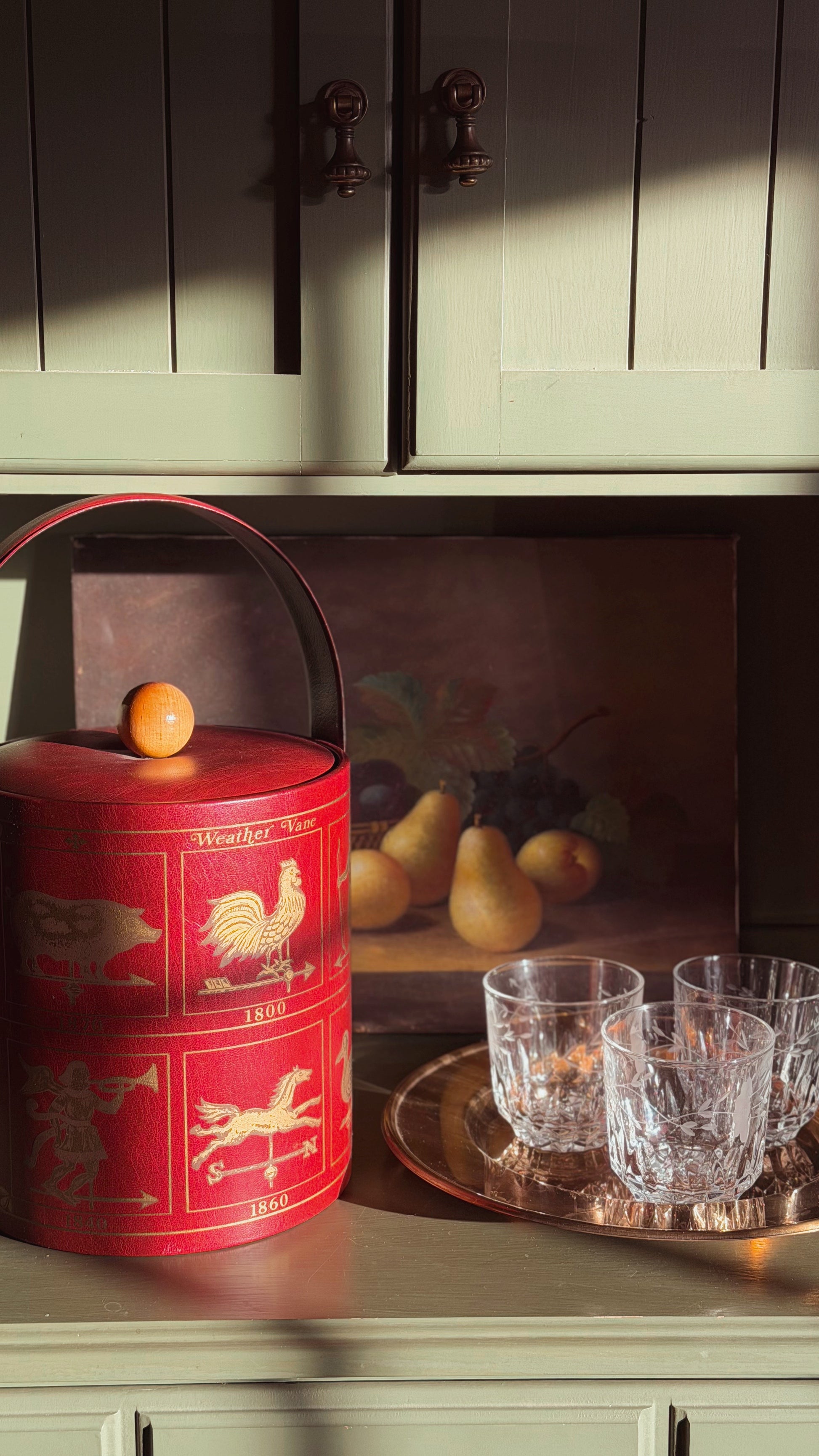 Vintage Georges Briard Weathervane Ice Bucket, placed on a cabinet next to clear cut glass tumblers on a tray in a warmly lit interior setting.