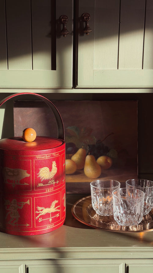 Vintage Georges Briard Weathervane Ice Bucket, placed on a cabinet next to clear cut glass tumblers on a tray in a warmly lit interior setting.