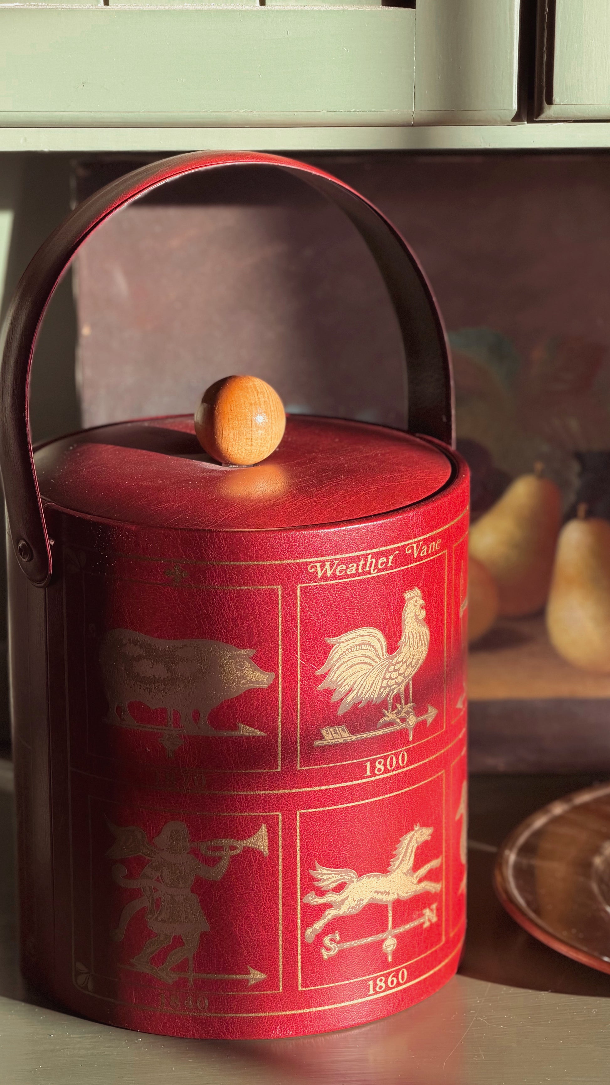 A red cylindrical lidded container with a wooden handle and knob, decorated with gold weatherman-style animal and figure illustrations, photographed on a shelf in warm light.