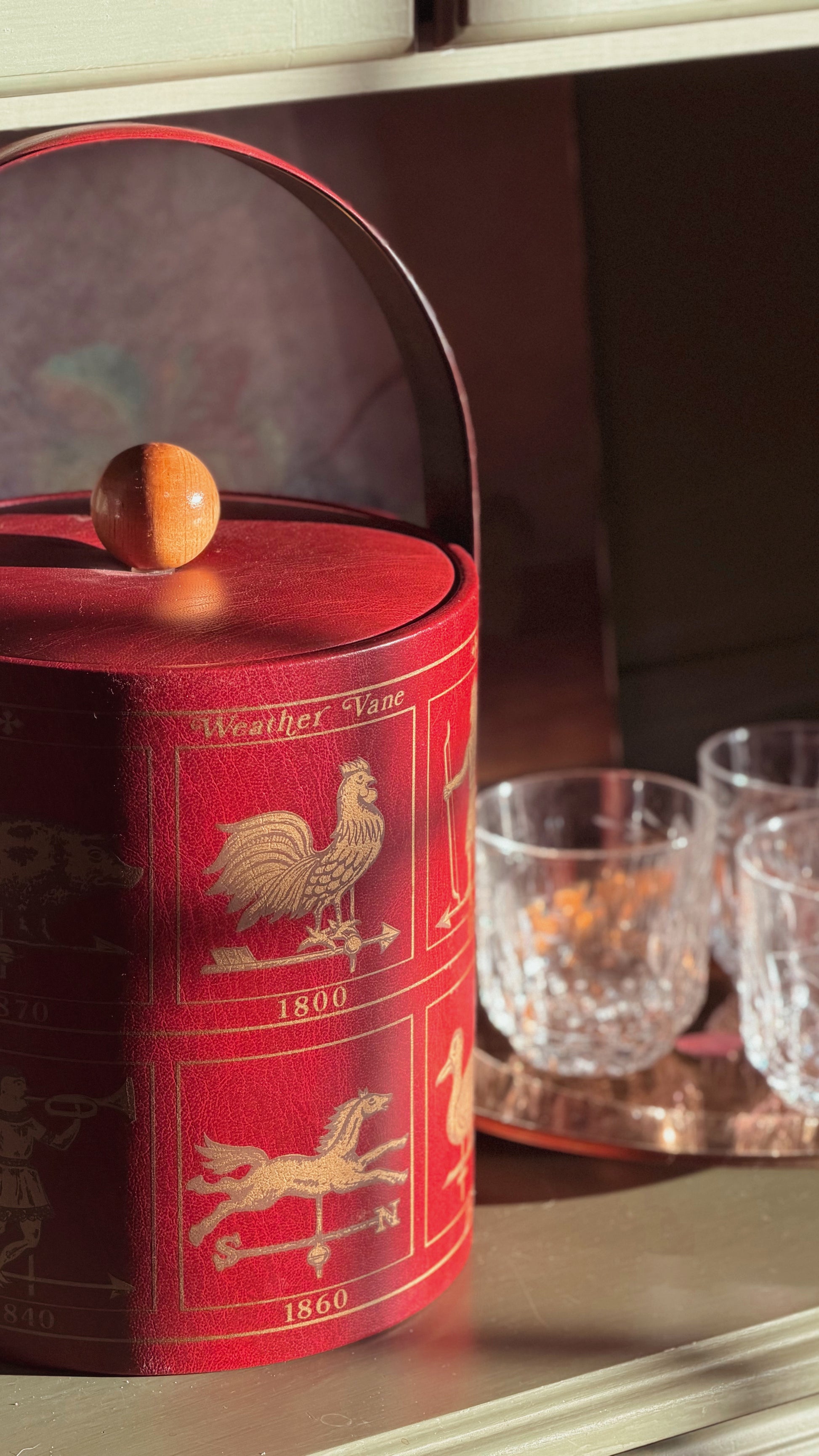 A red cylindrical weather vane tin with a wooden handle and knob, decorated with gold illustrations of weather symbols and years, shown on a shelf next to two clear drinking glasses.