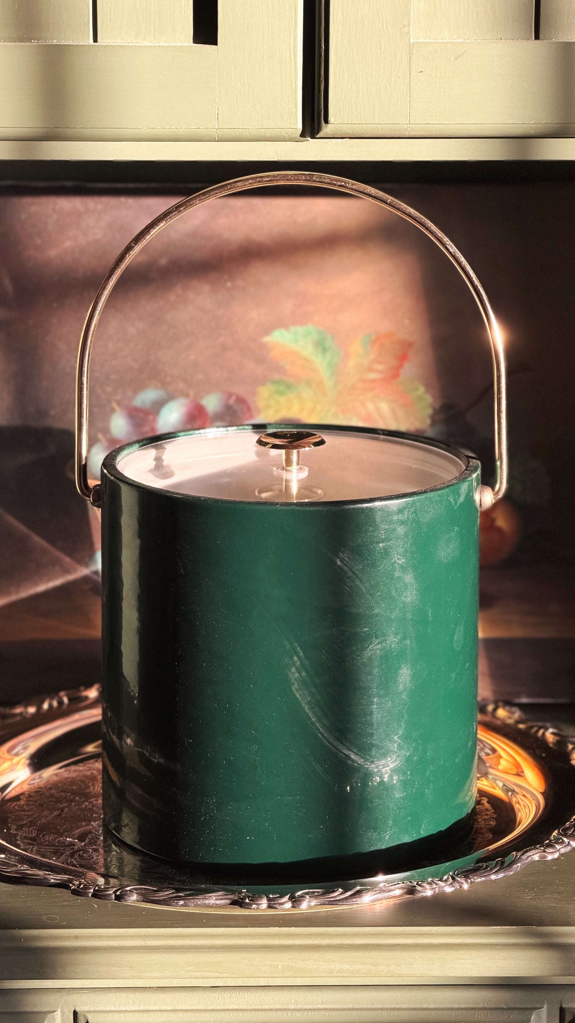A vintage green ice bucket with a clear glass lid and a curved metal handle, resting on a silver tray in warm, direct sunlight.