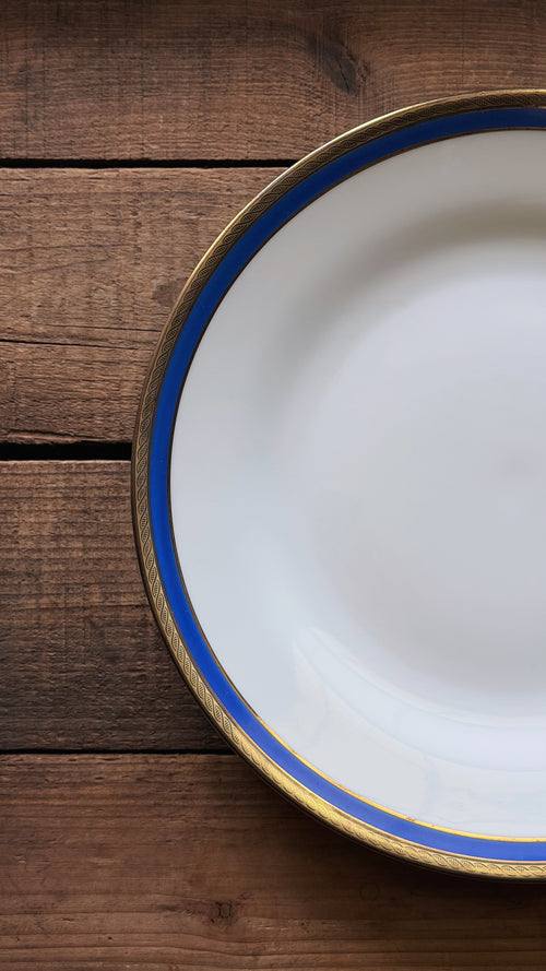 A Vintage Richard Ginori Palermo Blue Dinner Plate with a blue stripe and gold rim, shown on a rustic dark wood background.