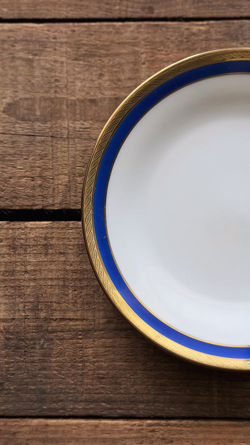 A close-up of a title: Vintage Richard Ginori Palermo Blue Bread & Butter Plate  with a blue and gold rim, placed on a rustic dark wood background.