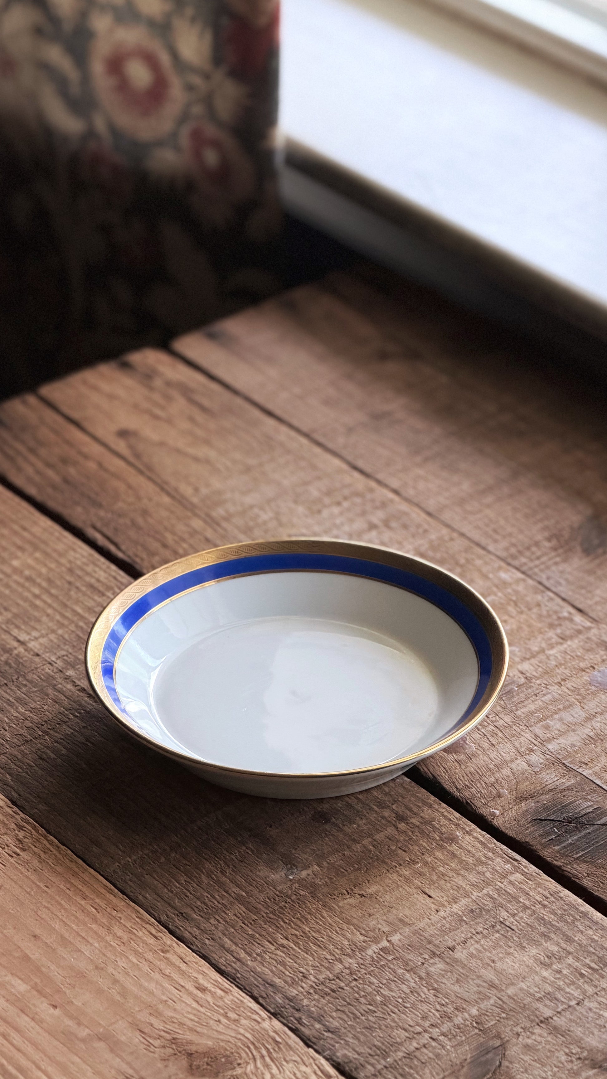 A Vintage Richard Ginori Palermo Blue Fruit Bowl with a blue and gold rim, resting on a rustic wooden table near a window.