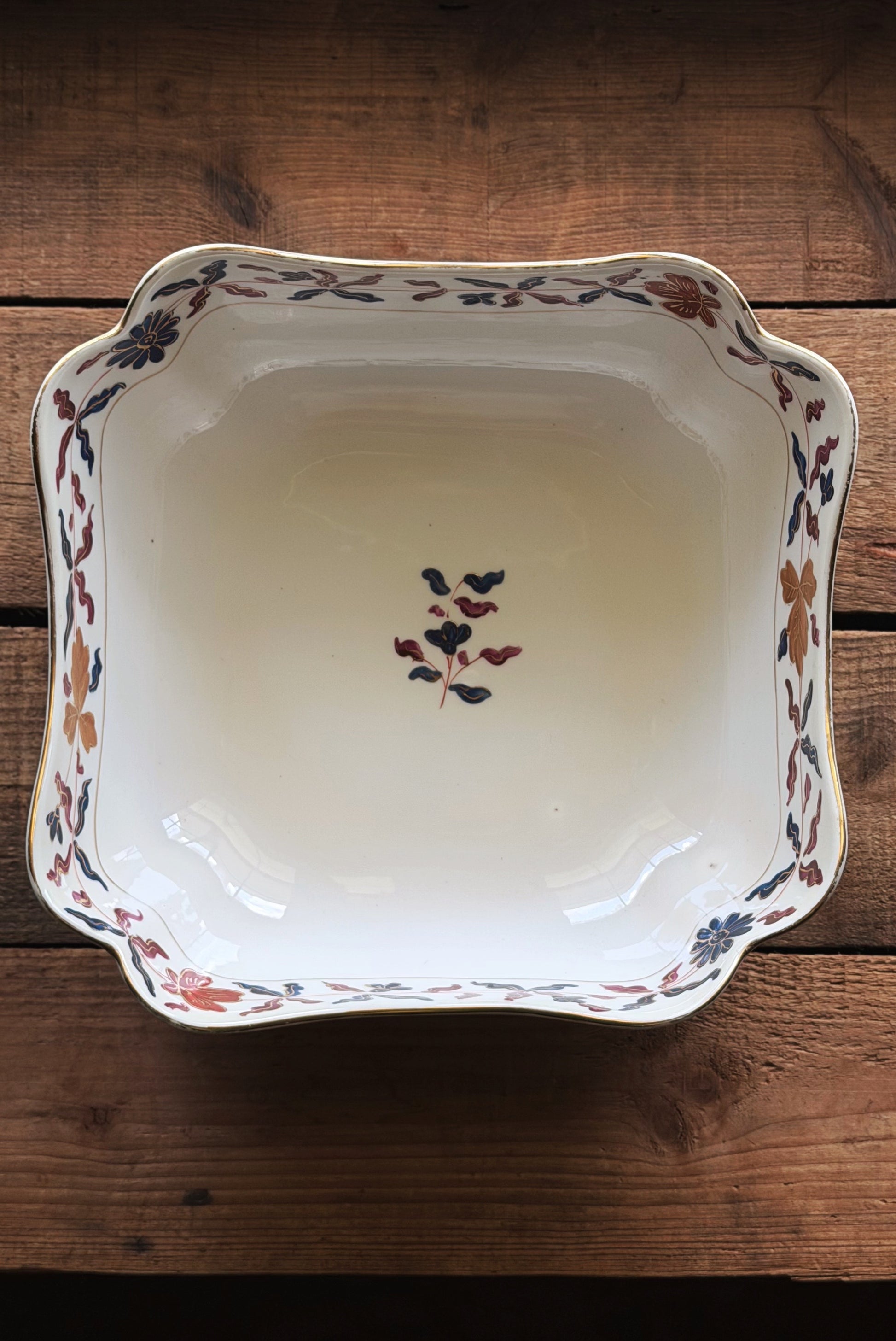 A square Vintage Royal Vienna Style Hand Painted Serving Bowl with scalloped edges, featuring a hand-painted floral pattern in muted red, blue, and brown on a white glaze, set against a rustic wooden background.