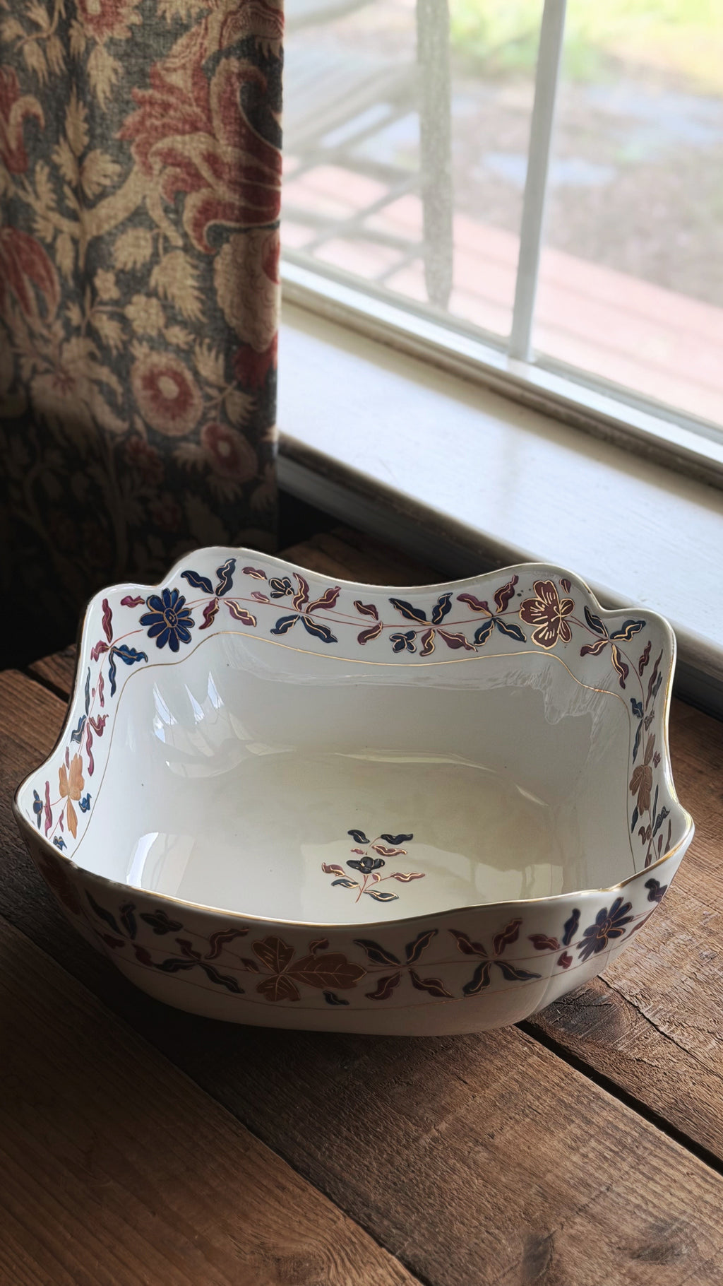 A Vintage Royal Vienna Style Hand Painted Serving Bowl with a scalloped rim and a delicate floral pattern in blue, orange, and brown, resting on a rustic wooden table next to a window.