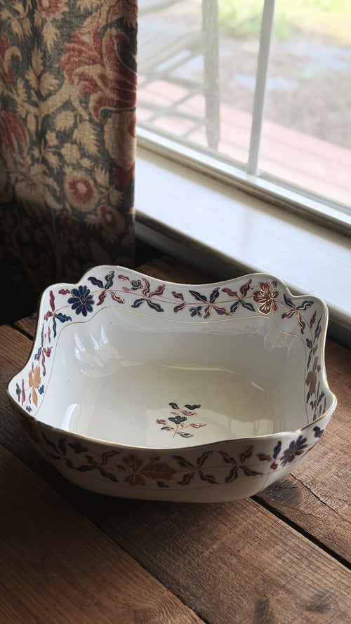 A Vintage Royal Vienna Style Hand Painted Serving Bowl with a scalloped rim and a delicate floral pattern in blue, orange, and brown, resting on a rustic wooden table next to a window.