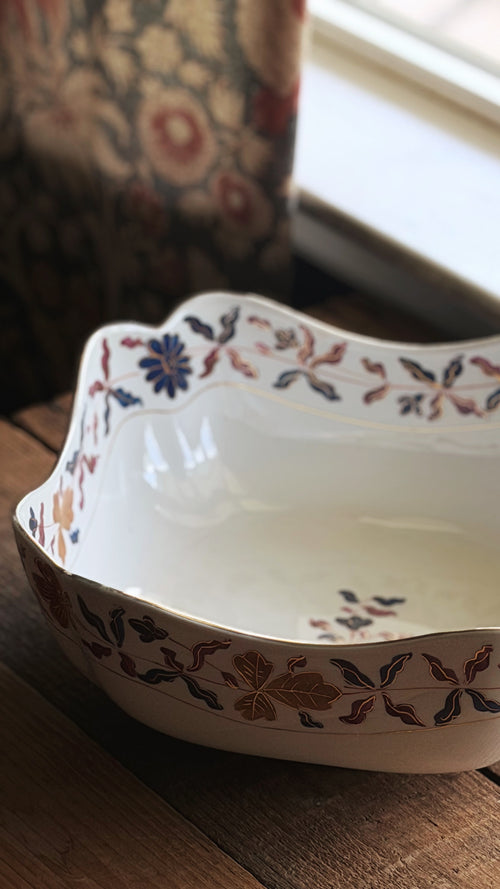 A Vintage Royal Vienna Style Hand Painted Serving Bowl with scalloped edges and a delicate floral pattern in blue, red, and gold, resting on a wooden table near a window.