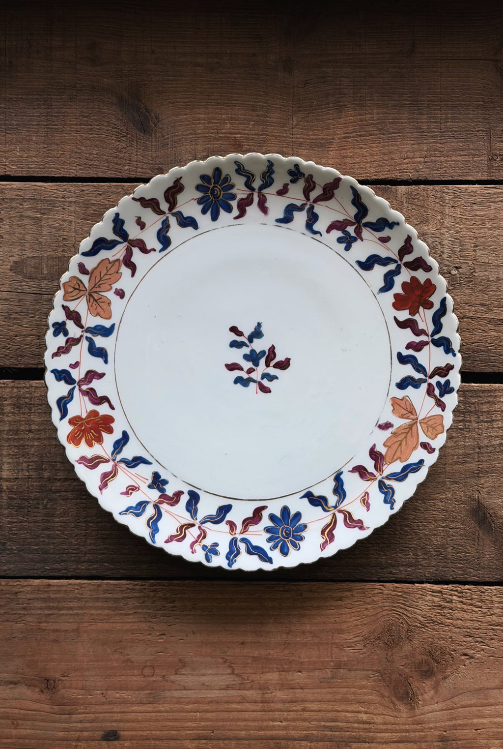 A Vintage Royal Vienna Hand Painted Salad Plate with a scalloped rim, decorated with a hand-painted floral and leaf pattern in blue, red, and orange, displayed on a rustic wooden surface.