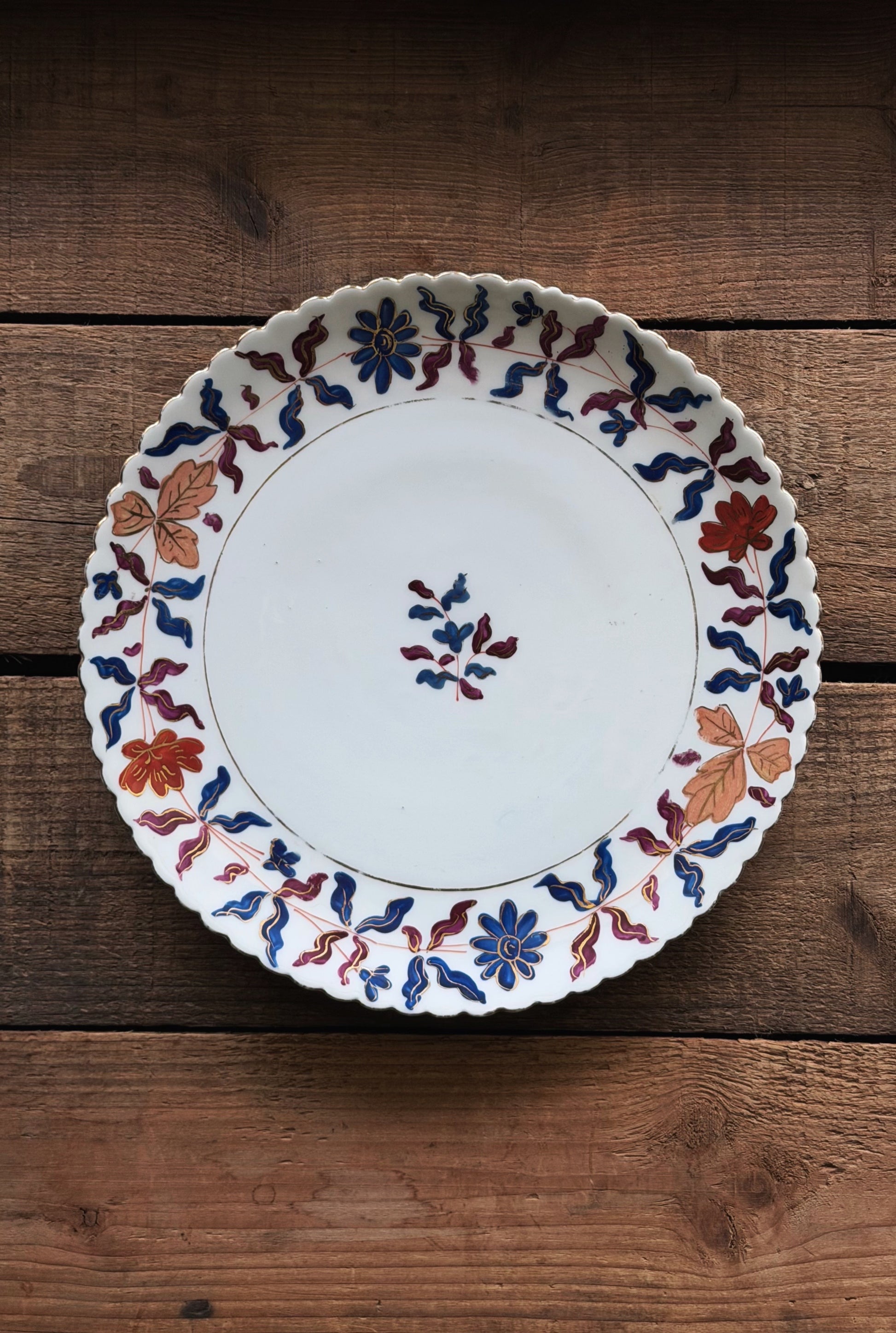 A Vintage Royal Vienna Hand Painted Salad Plate with a scalloped rim, decorated with a hand-painted floral and leaf pattern in blue, red, and orange, displayed on a rustic wooden surface.