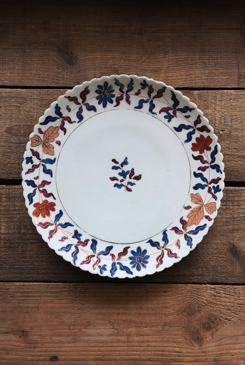 A Vintage Royal Vienna Hand Painted Salad Plate with a scalloped rim, decorated with a hand-painted floral and leaf pattern in blue, red, and orange, displayed on a rustic wooden surface.
