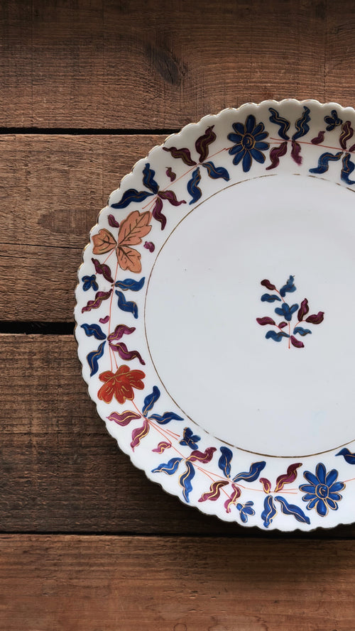 A Vintage Royal Vienna Hand Painted Salad Plate with a scalloped rim, decorated with blue, red, and orange floral and leaf patterns, resting on a rustic dark wood background.