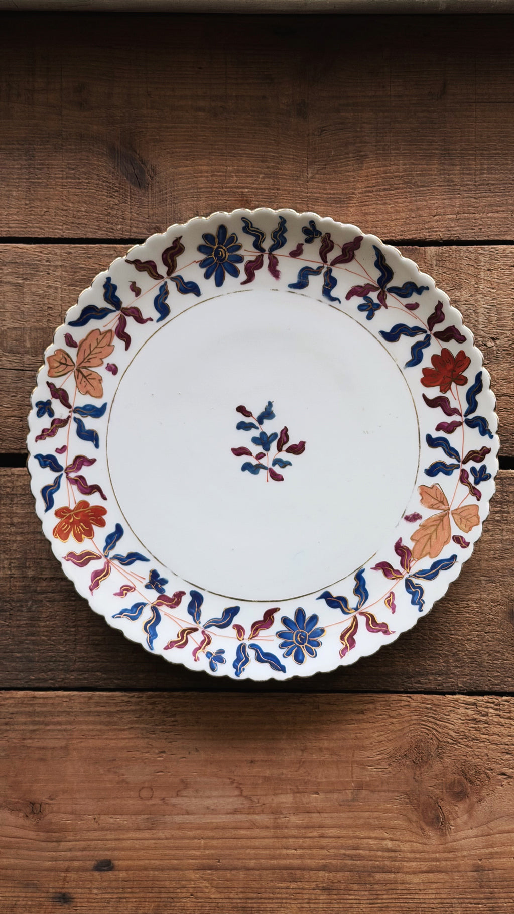 A round white ceramic plate with a scalloped rim, decorated with blue, red, and orange floral and leaf patterns, displayed on a rustic wooden surface.