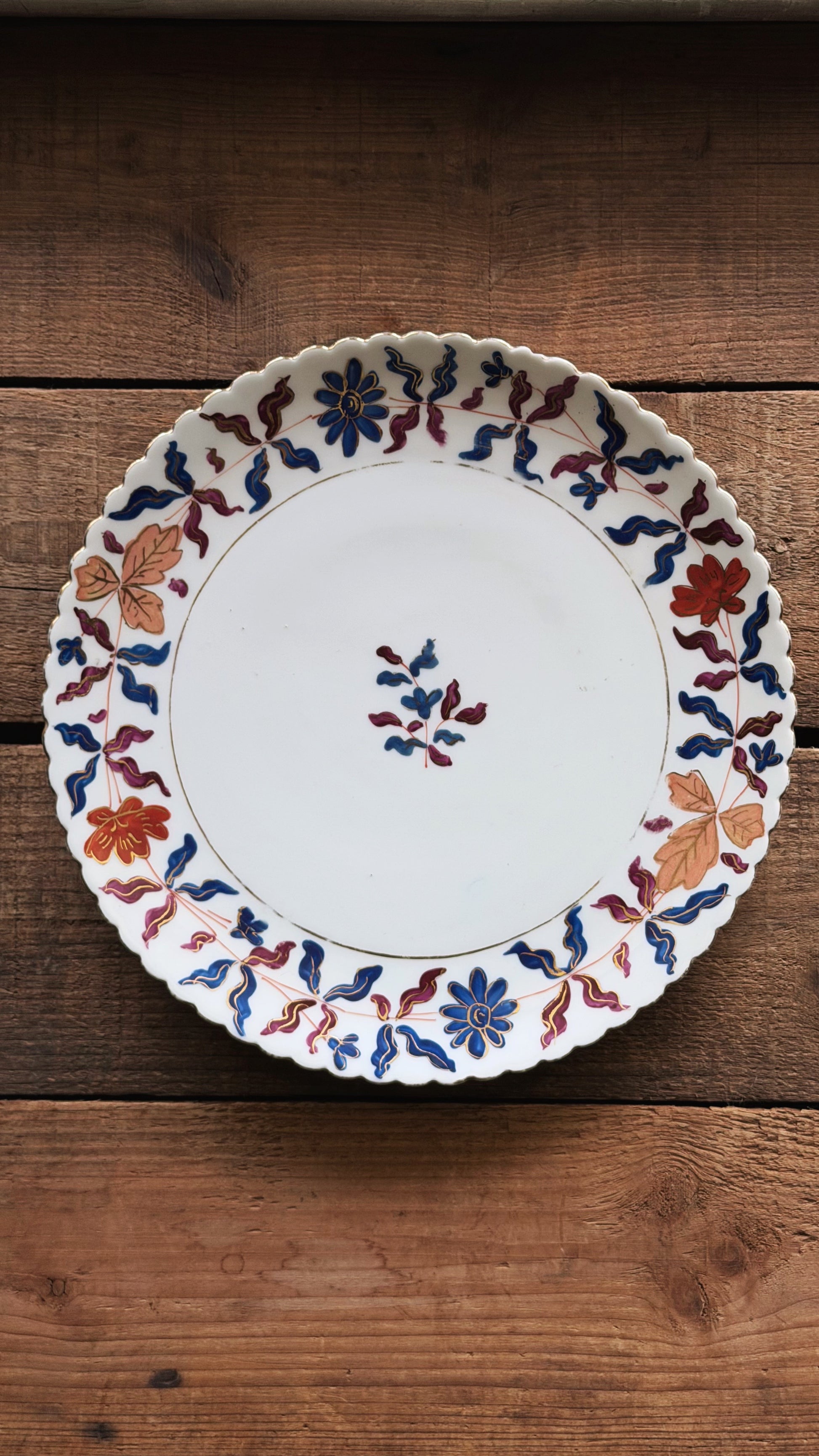 A round white ceramic plate with a scalloped rim, decorated with blue, red, and orange floral and leaf patterns, displayed on a rustic wooden surface.