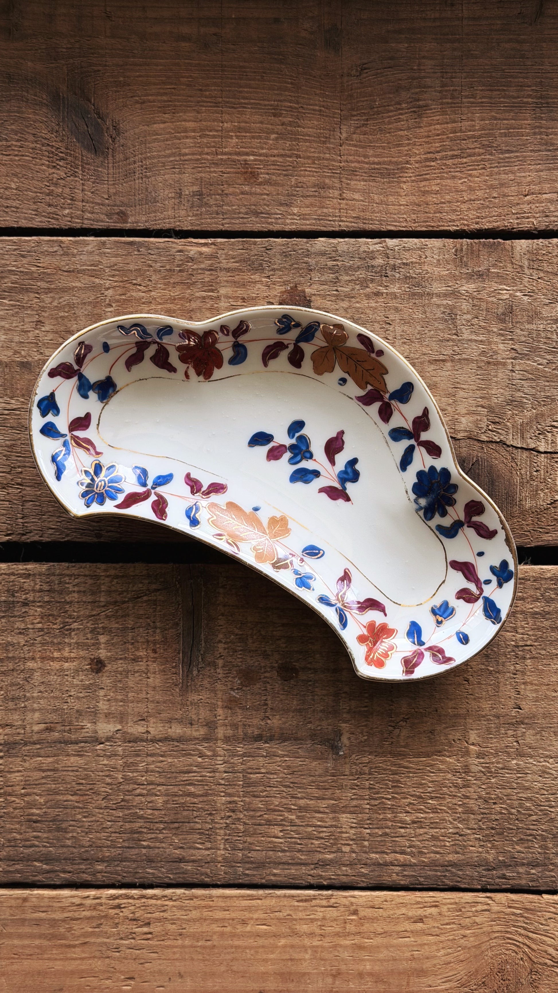 A Vintage Royal Vienna Hand Painted Bone Dish with a wavy, organic shape, featuring a white base decorated with blue, red, and orange floral motifs, set on a rustic wooden surface.