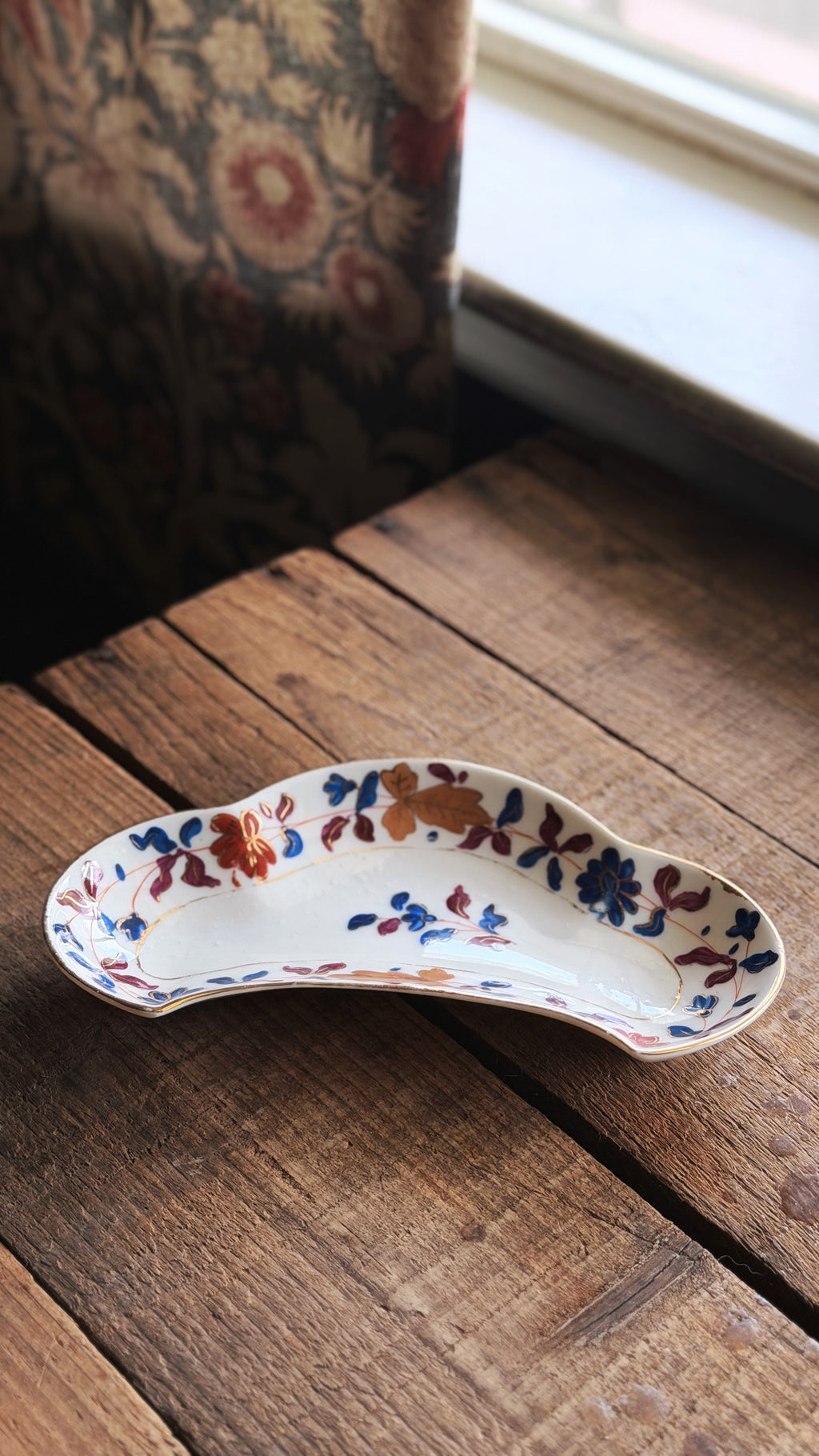 A Vintage Royal Vienna Hand Painted Bone Dish with a floral pattern in blue, orange, and red on a white base, resting on a rustic wooden table near a window.