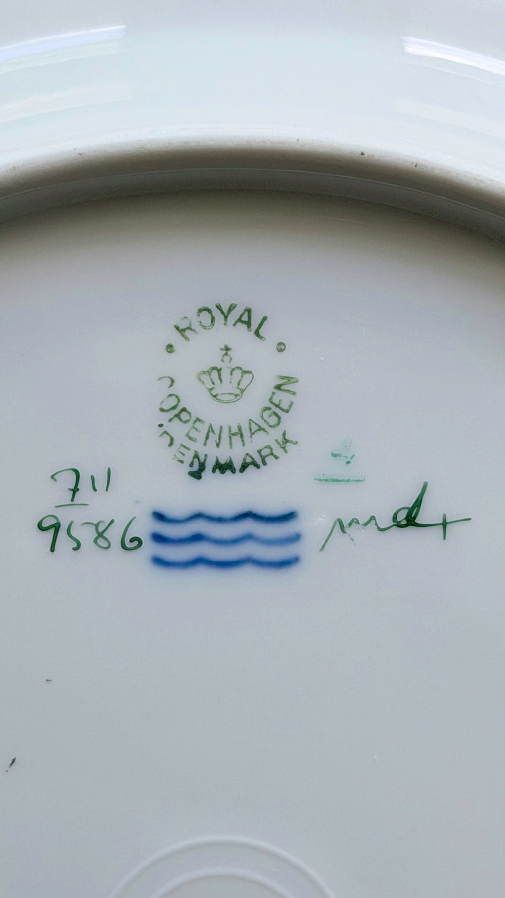 Close-up of the inside bottom of a white ceramic plate or dish, showing a green 'Royal Copenhagen' mark with a crown, blue wavy line decoration, and handwritten green and blue markings.