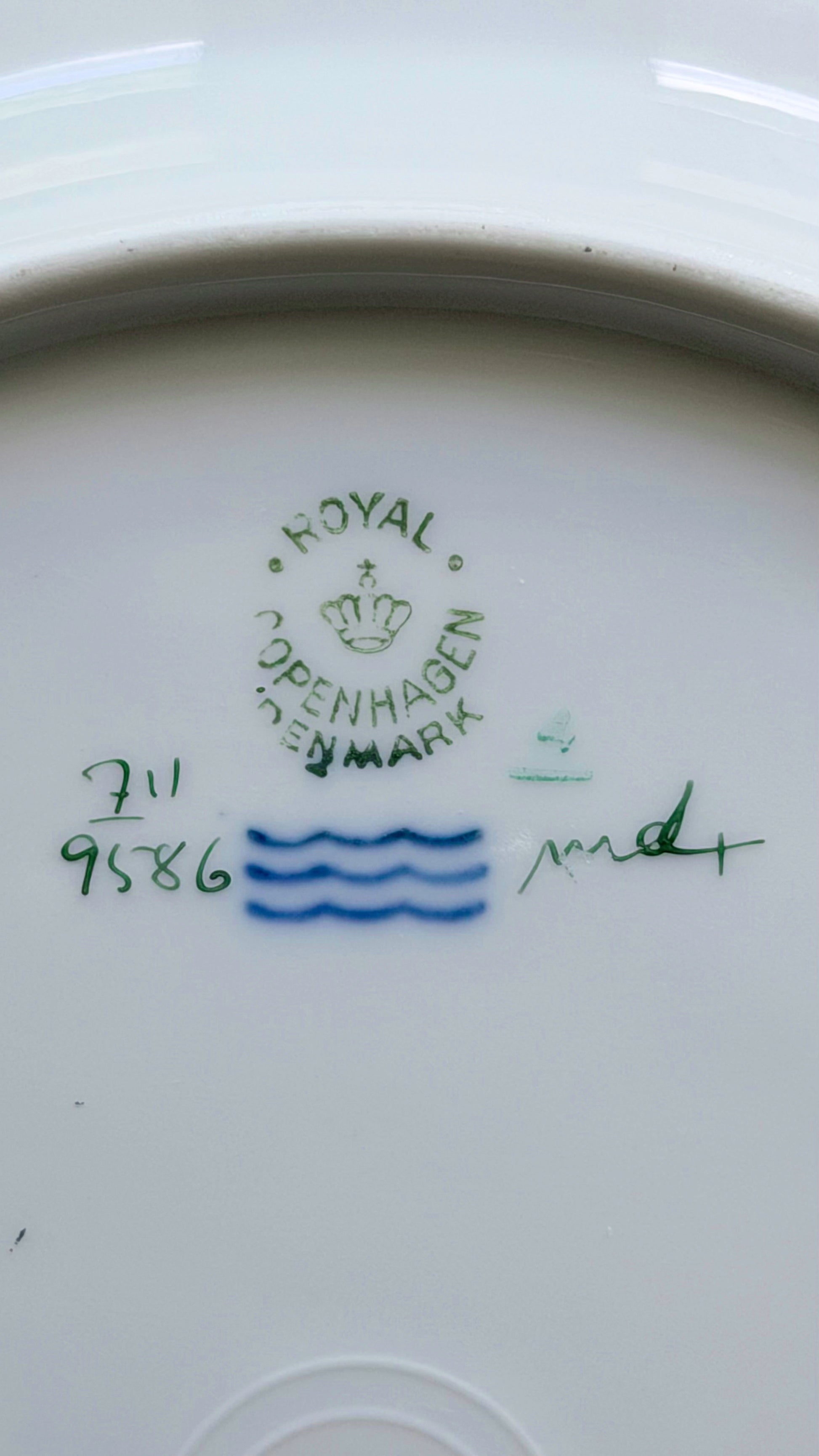 Close-up of the inside bottom of a white ceramic plate or dish, showing a green 'Royal Copenhagen' mark with a crown, blue wavy line decoration, and handwritten green and blue markings.