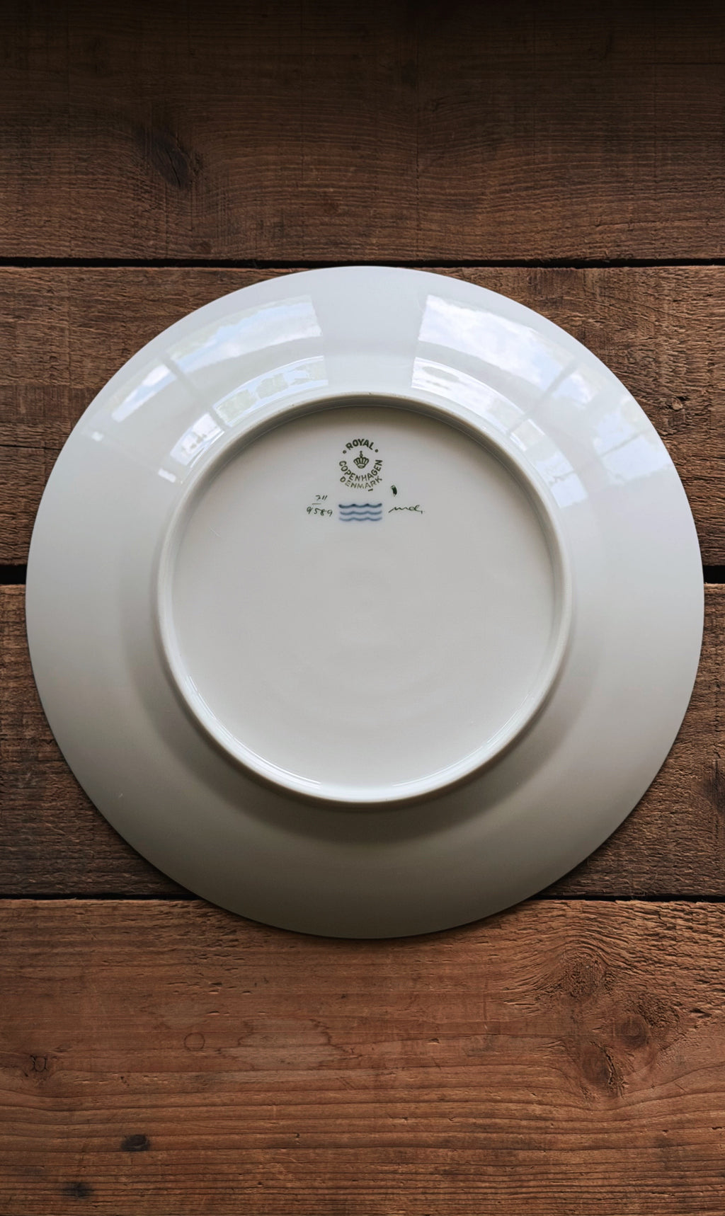 The back of a Vintage Royal Copenhagen Brown Iris Luncheon Plate with a small blue and green printed logo, placed on a rustic dark brown wooden surface.