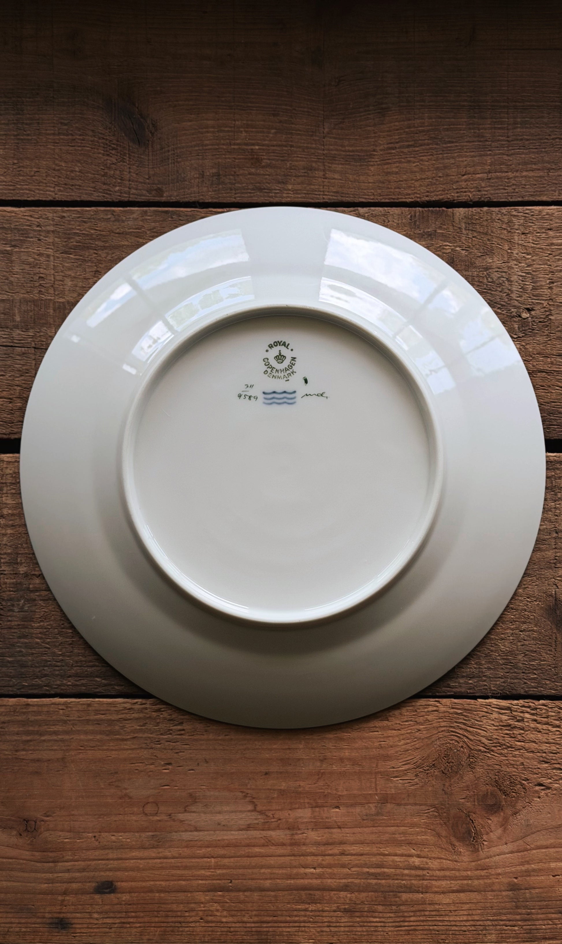 The back of a Vintage Royal Copenhagen Brown Iris Luncheon Plate with a small blue and green printed logo, placed on a rustic dark brown wooden surface.